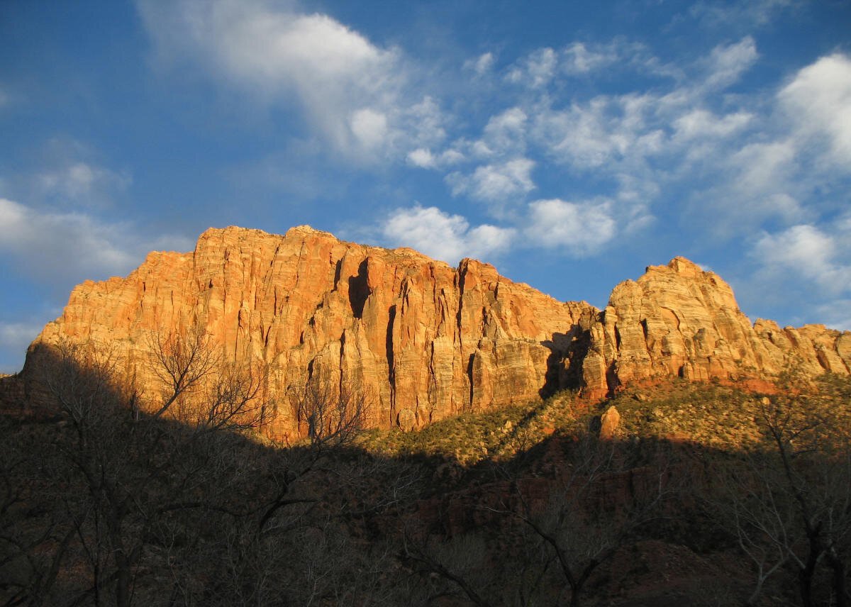A steep, rocky cliff is brightly literature by afternoon sun. Trees can be seen in the dark shadow of the foreground, and white, puffy clouds are scattered in the blue sky above.