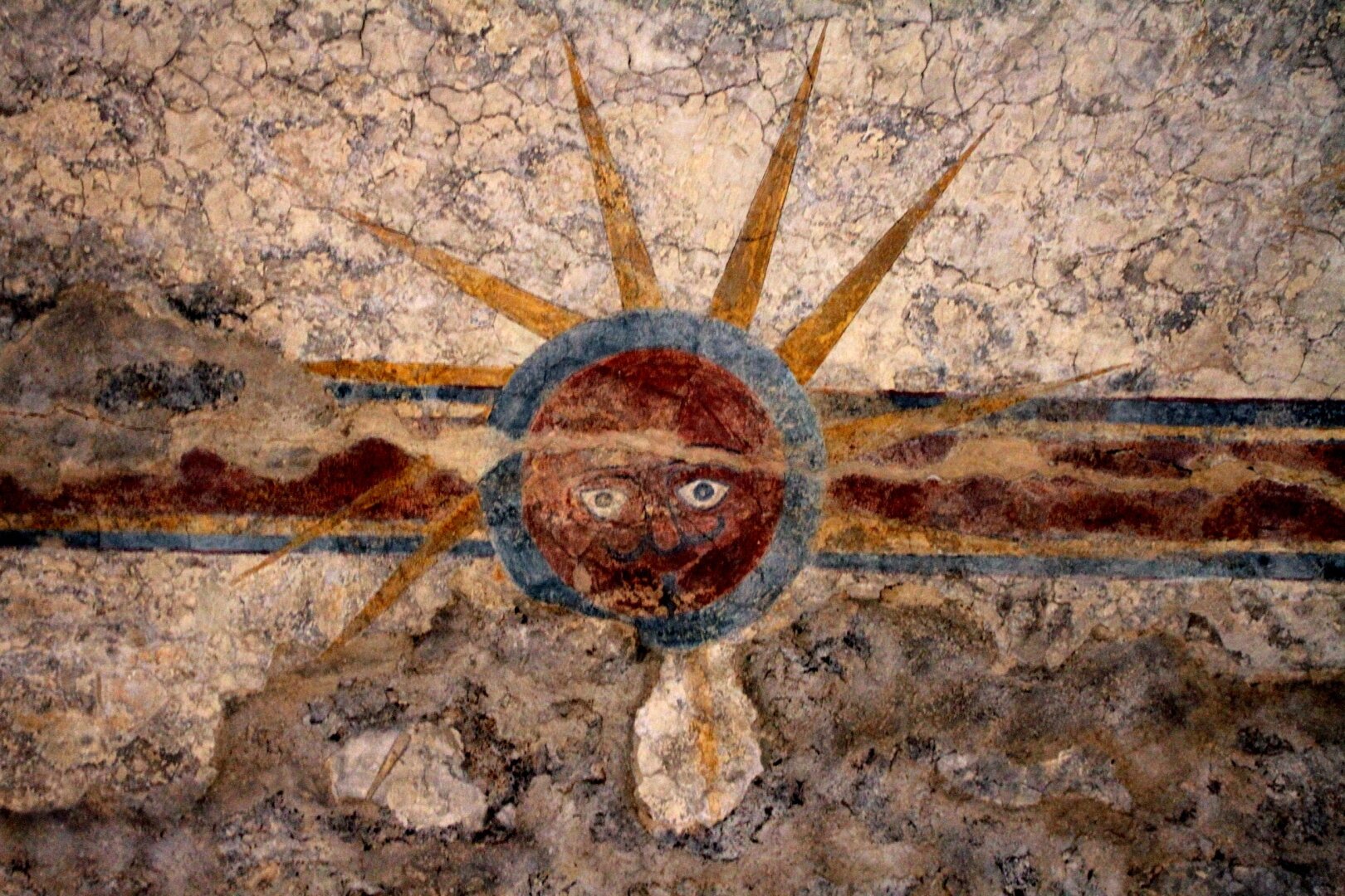 This ancient ceiling painting at Mission Concepción in San Antonio, Texas, depicts a sun with a human face. The red and brown visage features white eyes and radiating yellow sunbeams against a rough, textured stone background.