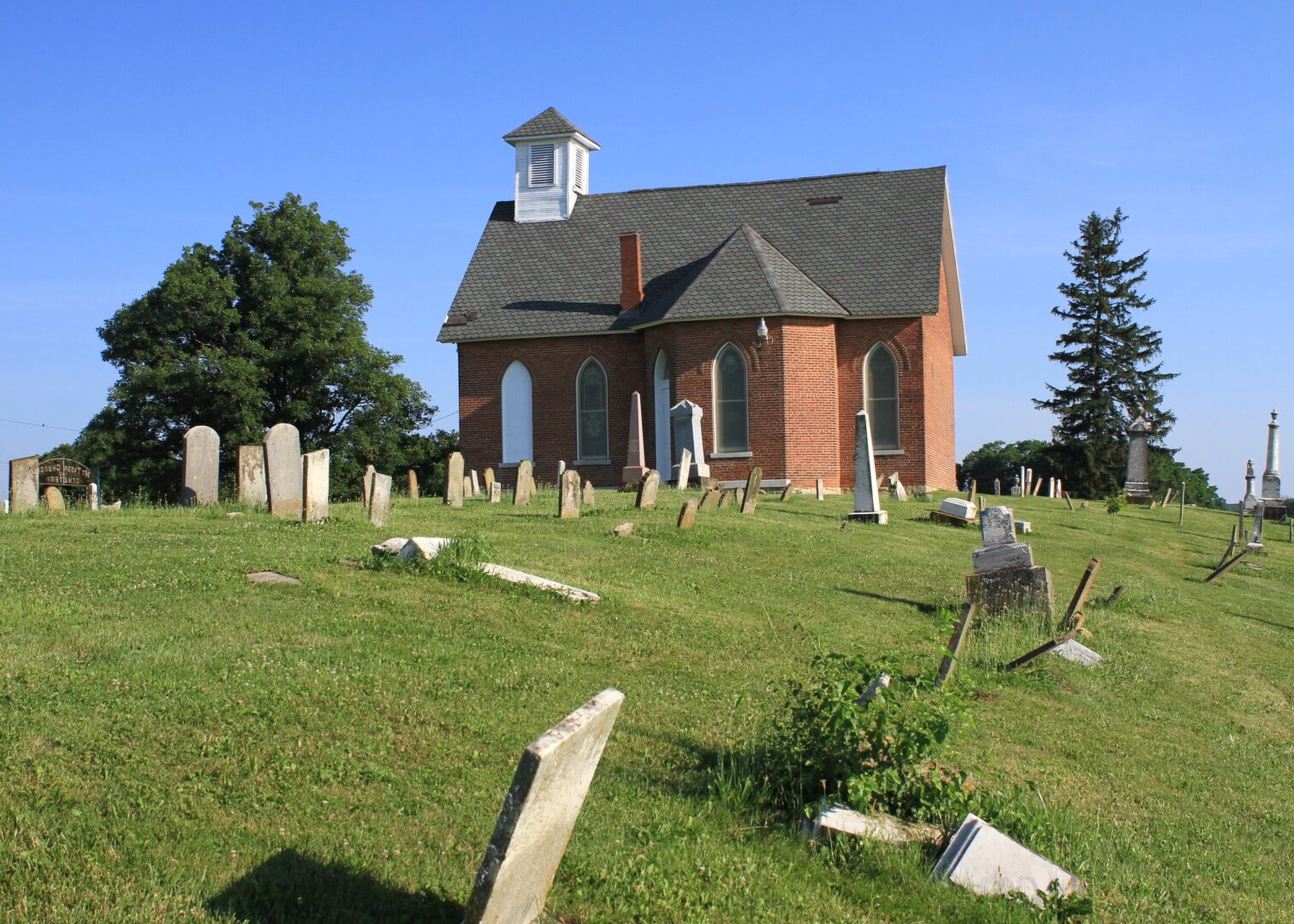 A small, red-brick church with a white steeple sits on a grassy hill surrounded by numerous old, leaning gravestones under a clear blue sky, with trees providing a serene backdrop.