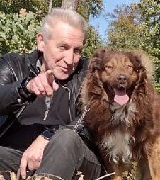 Das Foto zeigt links einen Mann in Lederjacke, der lächelnd auf den Betrachter deutet und rechts daneben einen sehr kaukasisch anmutenden Hund, der ebenfalls direkt in die Kamera zu lächeln scheint.