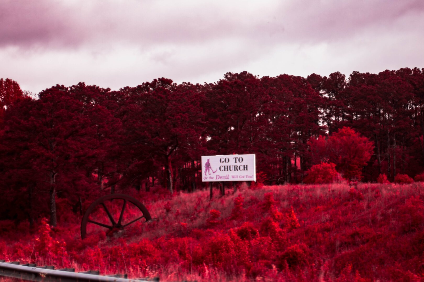 An odd scene, assign reads Go to church or the devil will get you, with the red figure of the devel with a scythe. To the left a large metal wheel is half buried and they are surrounded by red grass of field trees are A dark red ish brown in the background and behind them is a cloudy sky. The railing of a highway can be seen in the left bottom corner.