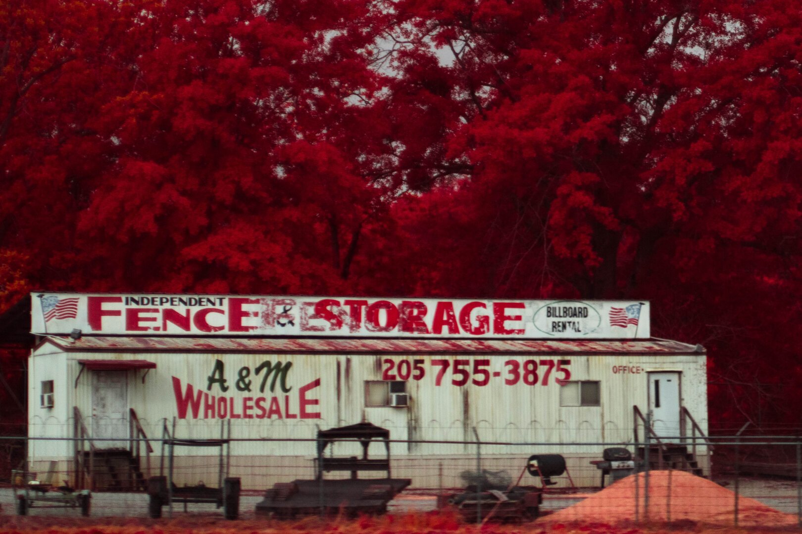 A mobile home turned business, this fence and storage business is surround by lose rusting machinery. Two dilapidated stares run up to two separate doors on the advertisement littered wall.