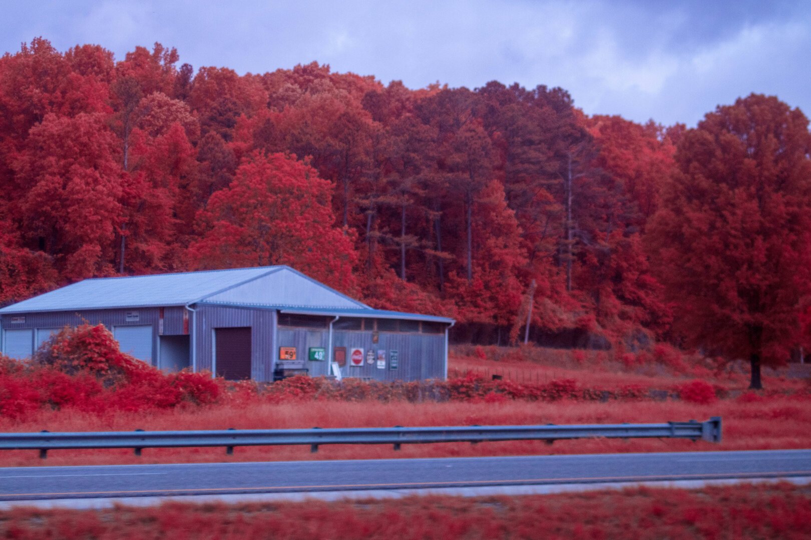 A small collection of sins hang on the wall of this barn house or shop. A small hill rises behind it and the interstate in front with a stop of guardrail to ward star cars from the building. The whole imagine is shifted red.