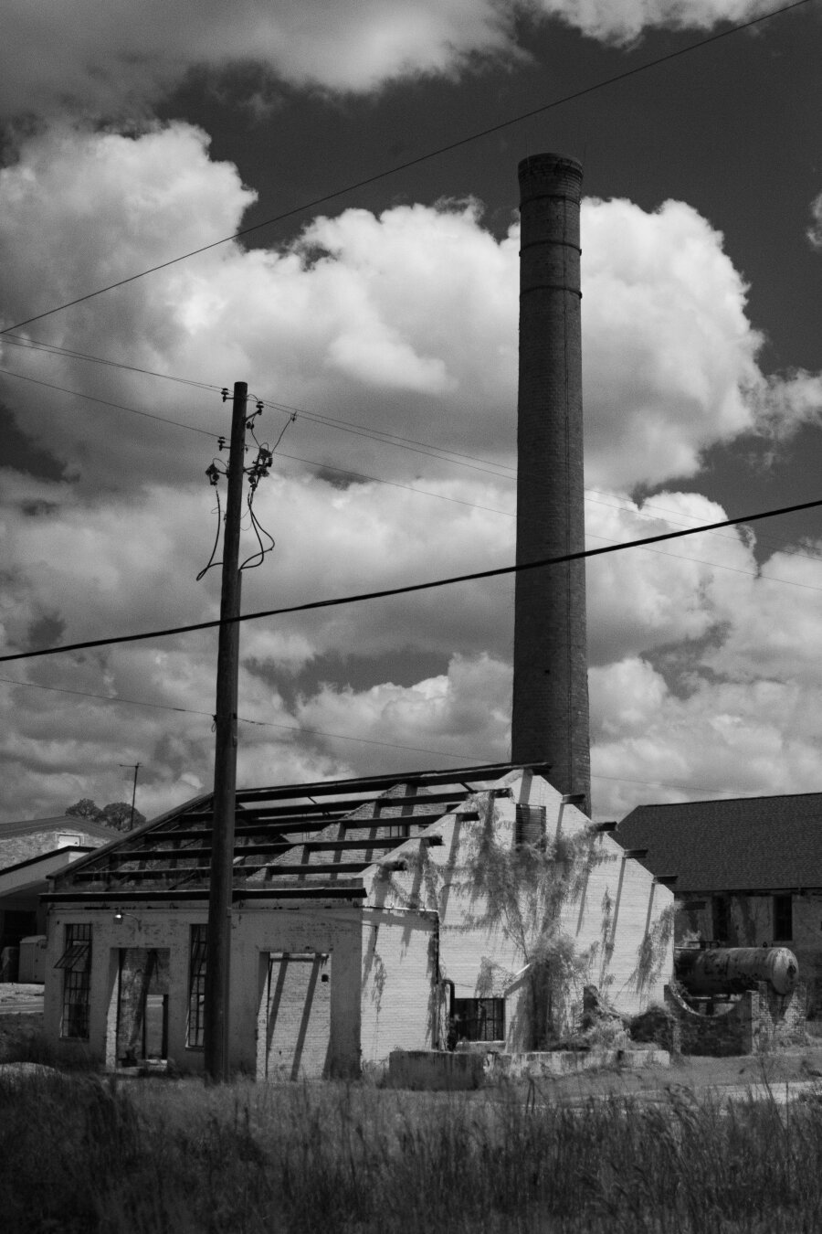 An old roofless structure sits between a telephone pole and a chimney stack stretching up into a hole in the cloudy sky. Moss drapes like tinsel from the bright sides where a small section stands a dilapidated propane tank.