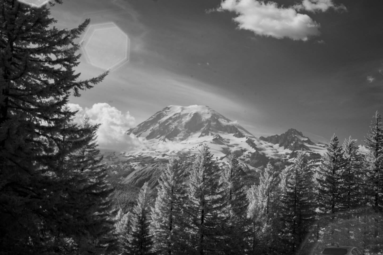 The snow capped rocky summit of Mt Rainier stands behind a line of foreground firs that dope to show the valley and rolling mountainside below a scarf of clouds around the snowline. Three sets of 7 sided flares cross the frame from the sun int the top left to the roof of a Ford mach-e on the bottom right. The sky is darkened dispite being in the dead of day because this photo was shot in light hight than 850nm, primarly the near infrared, leaving dramatic black, grays and whites.
