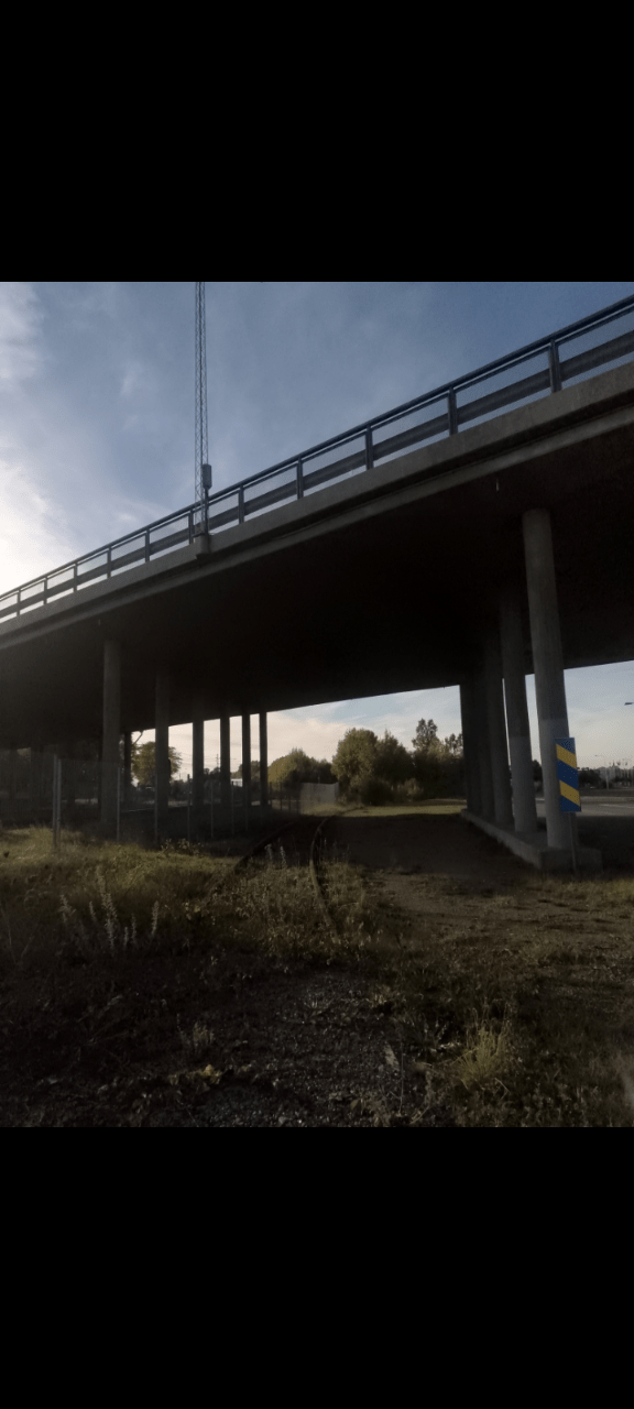 Part of an abandoned traintrack. On the left side is some columns, a fence and then a highway. On the right is also some columns and then a small road. The columns support a highway overpass.