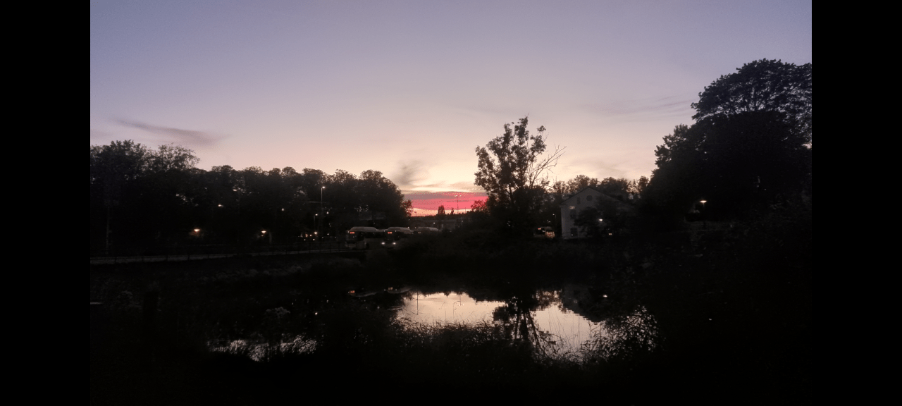 A picture of the sunset. Part of a busstation is visible. Theres also a small lake in the foreground. Some trees on both left and right side. A house the right of the busstation is visible and also a bus on the busstation.
