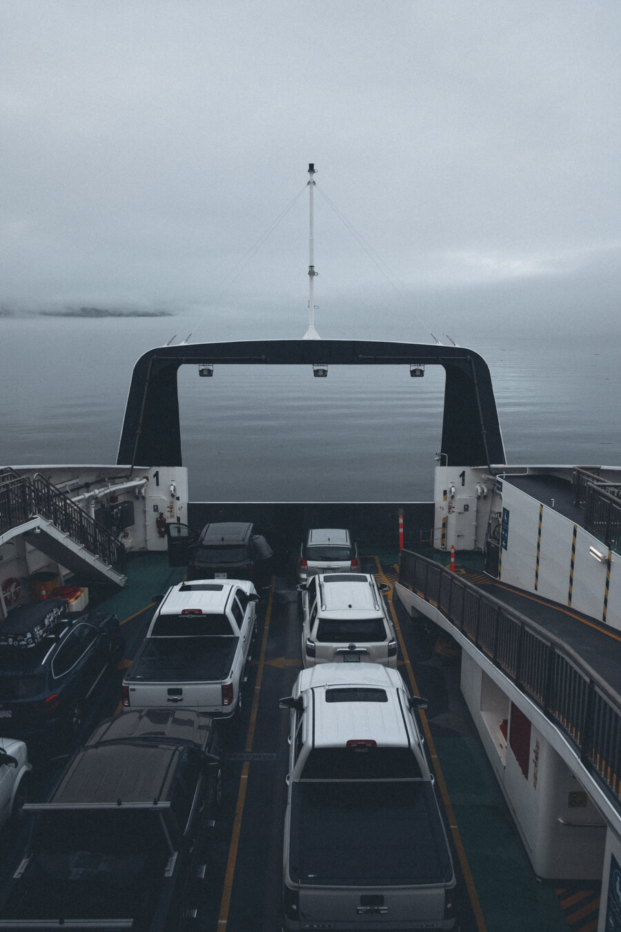 A cloudy, misty morning at sea as several cars and trucks sit parked on the deck of a ferry, with calm water and a distant shoreline in the background.