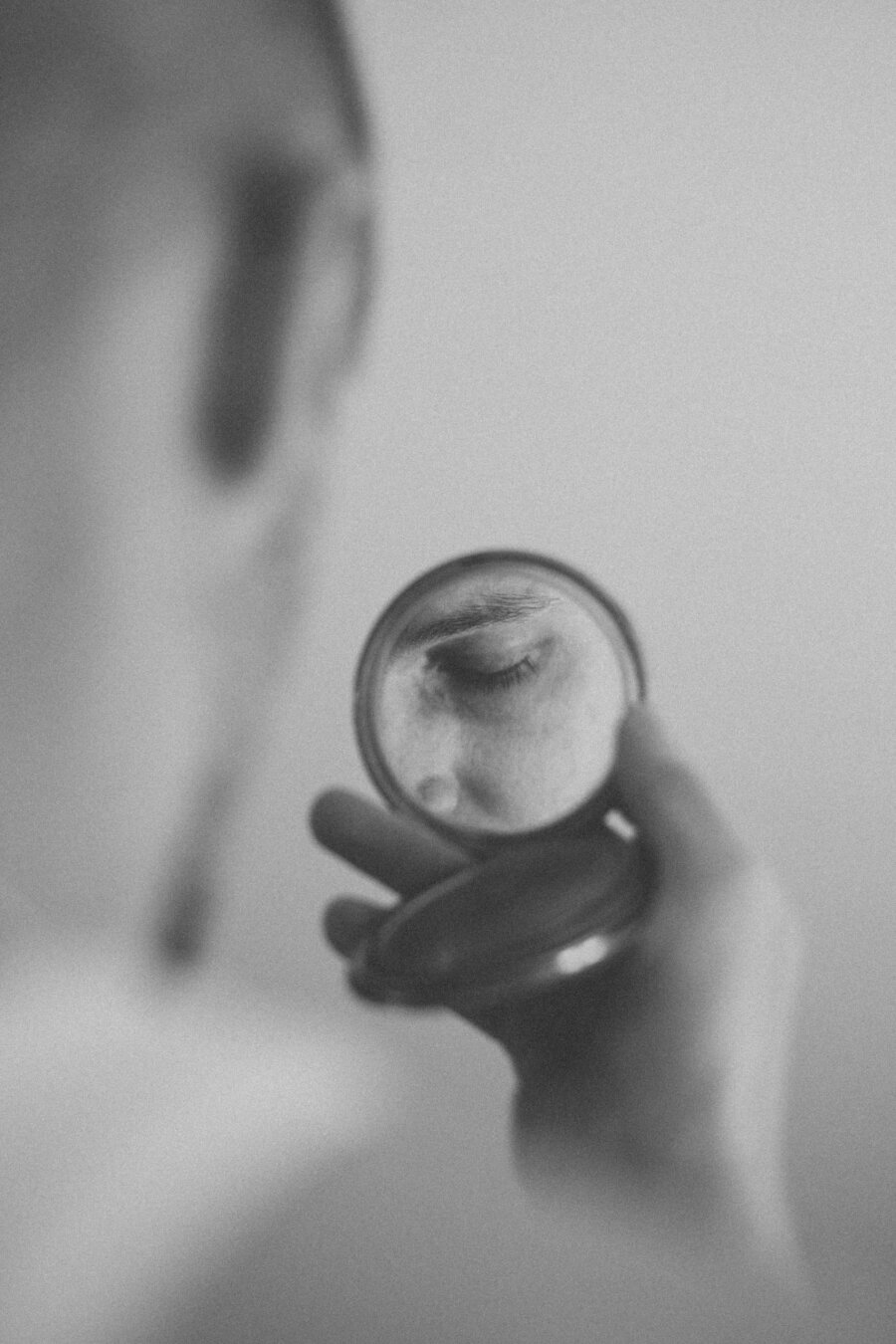A monochrome photo of a person holding a small round mirror that reflects their closed eye, creating a calm and introspective mood with a soft, blurred background.