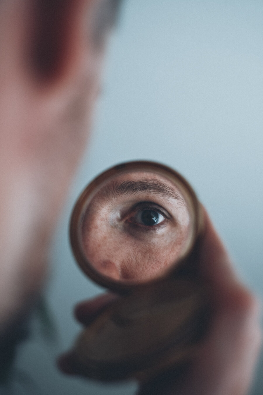 A close-up photograph showing a person looking through a small round mirror that reflects one of their eyes in sharp detail, while the rest of the image is softly blurred.