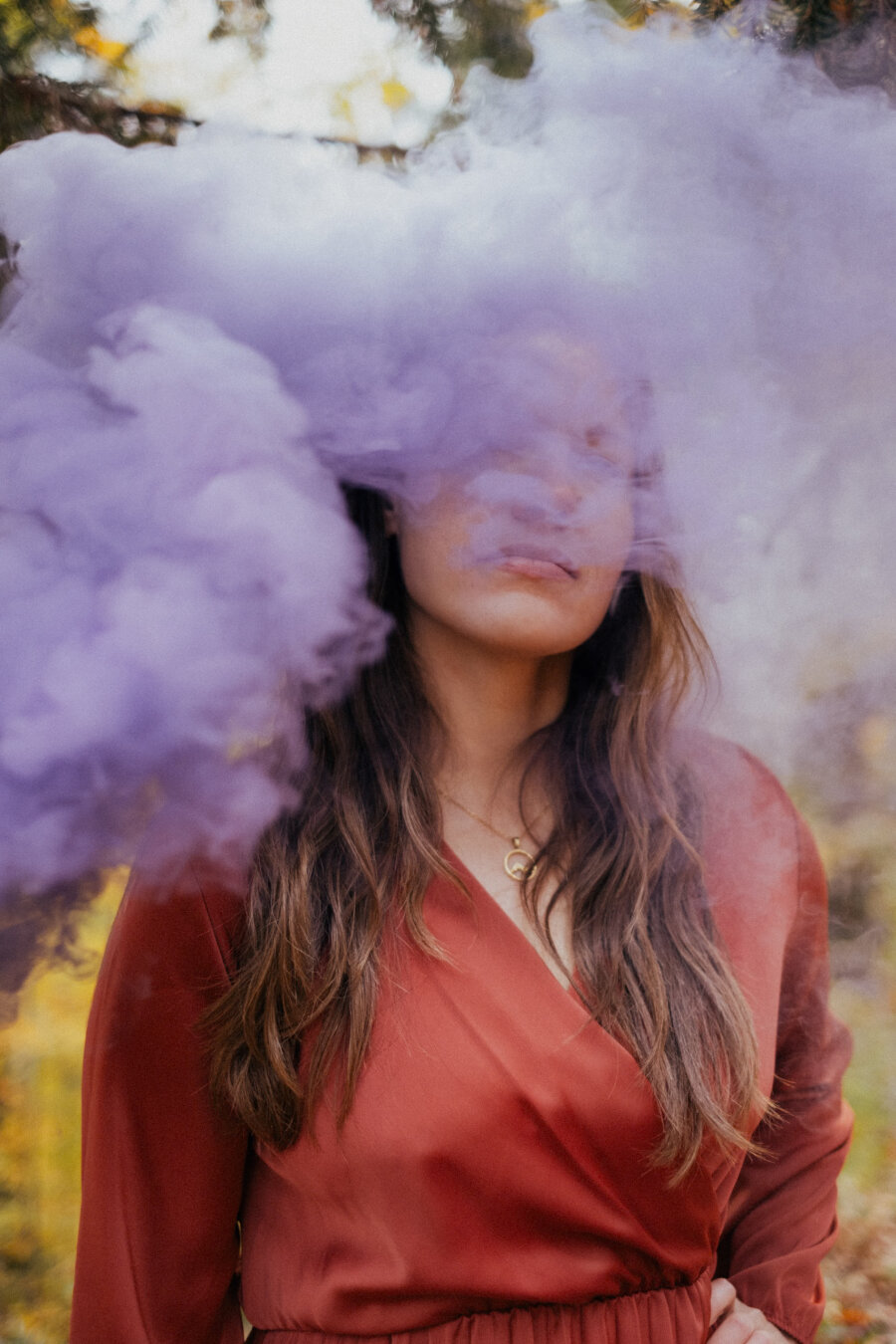 A person wearing a long-sleeved, rust-red dress stands outdoors while thick purple smoke drifts across their face, obscuring their eyes. Their long, wavy brown hair falls over their shoulders, and they wear a gold necklace. The background is softly blurred with hints of greenery and warm tones.