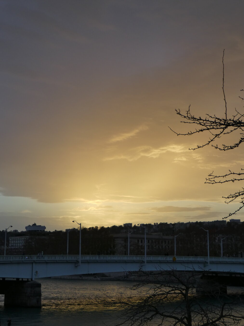 Picture of a bridge with buildings behind. The Sun has set behind the buildings, and its light keeps emanating from there. We can also distinguish some rays of light.