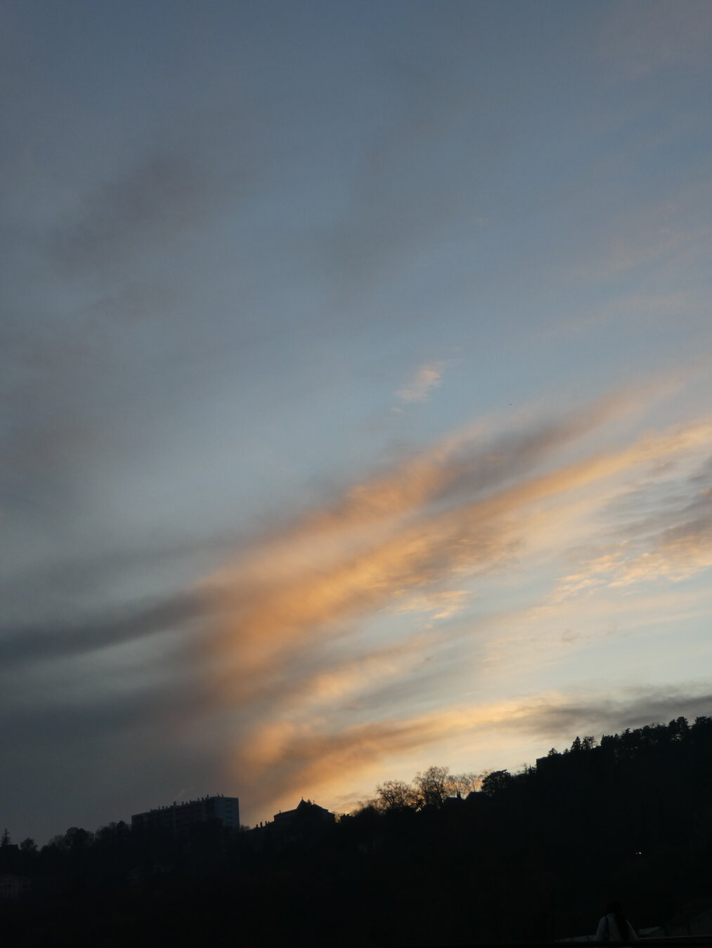 Picture showing the sky and some clouds illuminated by the light of a sunset. On the bottom we can see part of a hill with buildings on it.