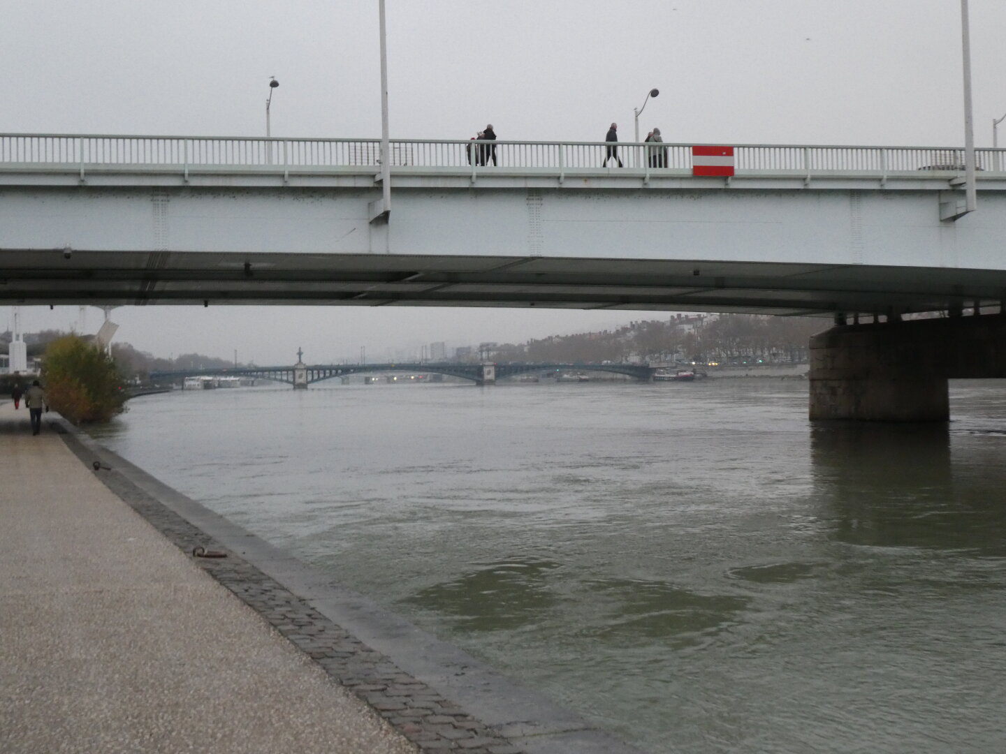Picture of a bridge crossing over a river. People can be seen walking on the bridge. On the left we can see the quay the picture was taken from, and some people walking on it too. Another bridge can be seen far away.