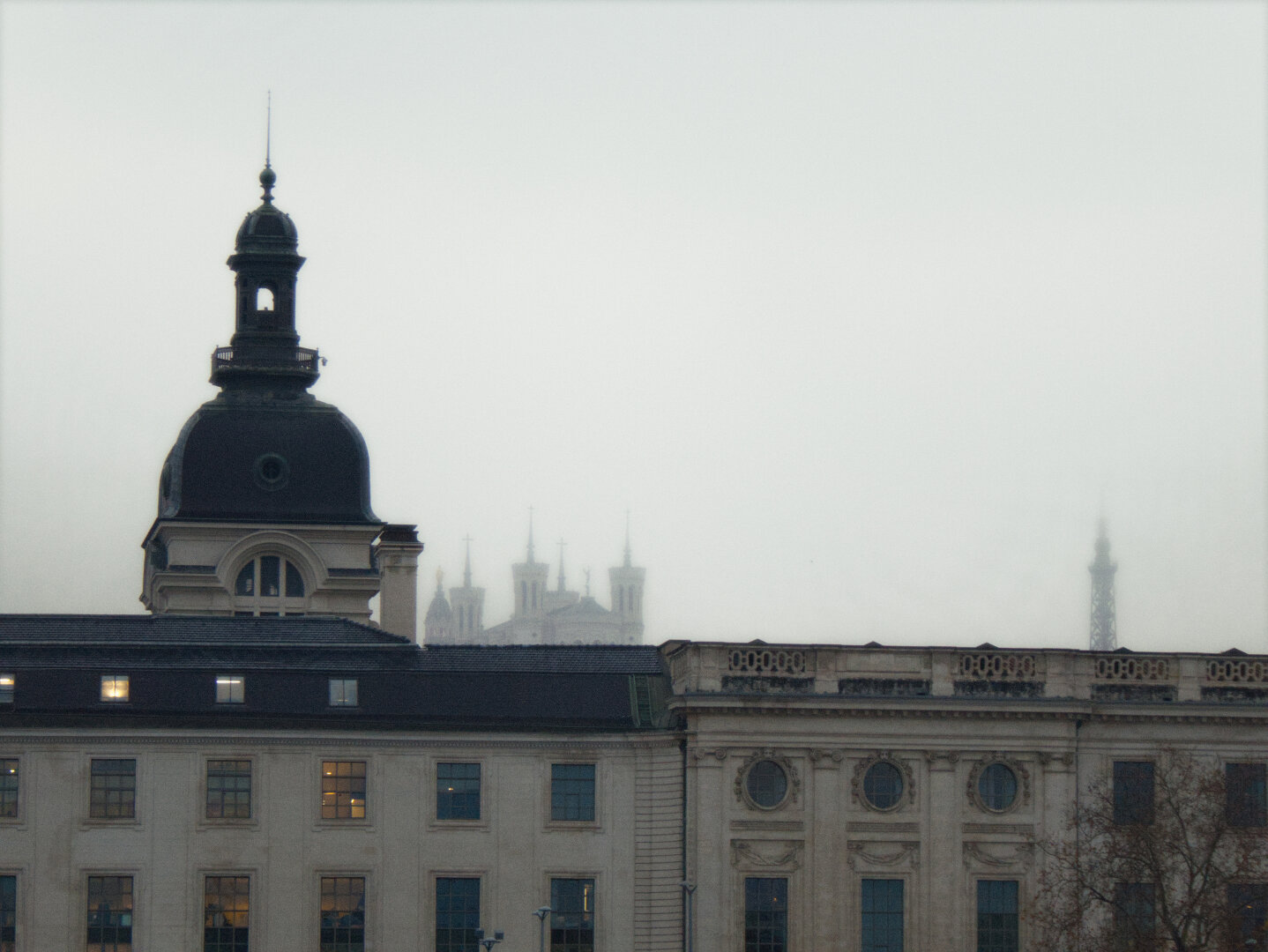 Picture of a french neo-classical building with a tower. From behind the building on the tower's right, a church can be seen. There's a replica of the Eiffel tower on the right side of the picture and from behind the building.