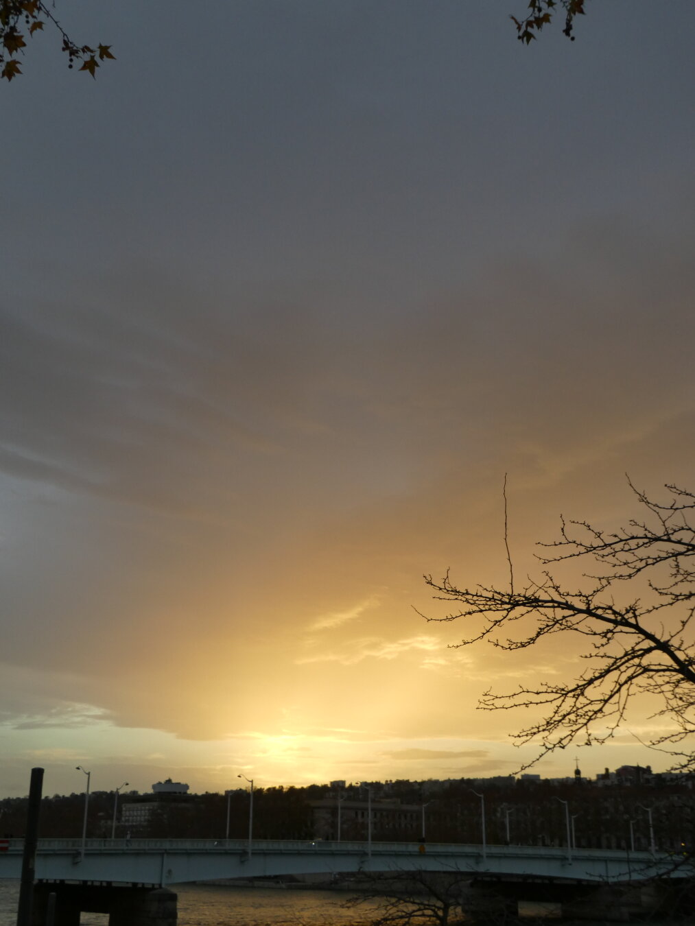 Picture of a bridge with buildings behind. The Sun has set behind the buildings, and its light keeps emanating from there.