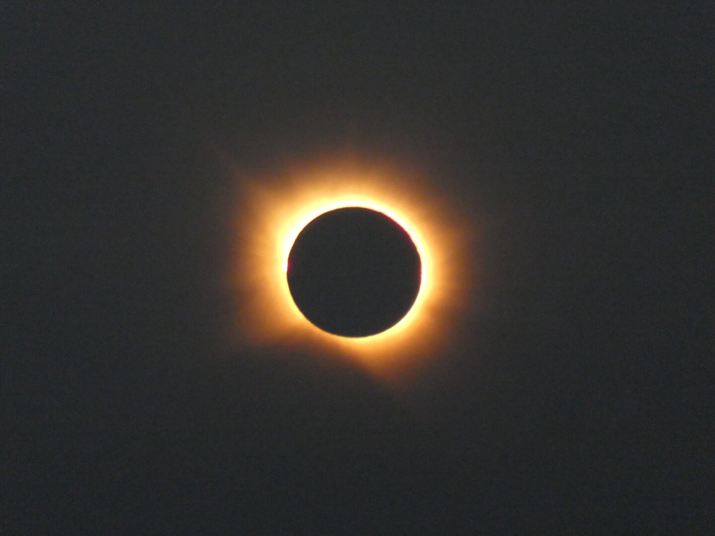 The moon eclipsing the sun in its entirety. Light from the sun can be seen shin all around the moon, lighting up an otherwise greyish black backdrop.
