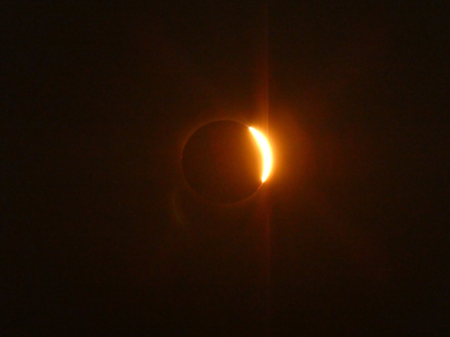 The moon eclipsing the sun in its entirety. Light from the sun can be seen shin all around the moon, lighting up an otherwise greyish black backdrop.