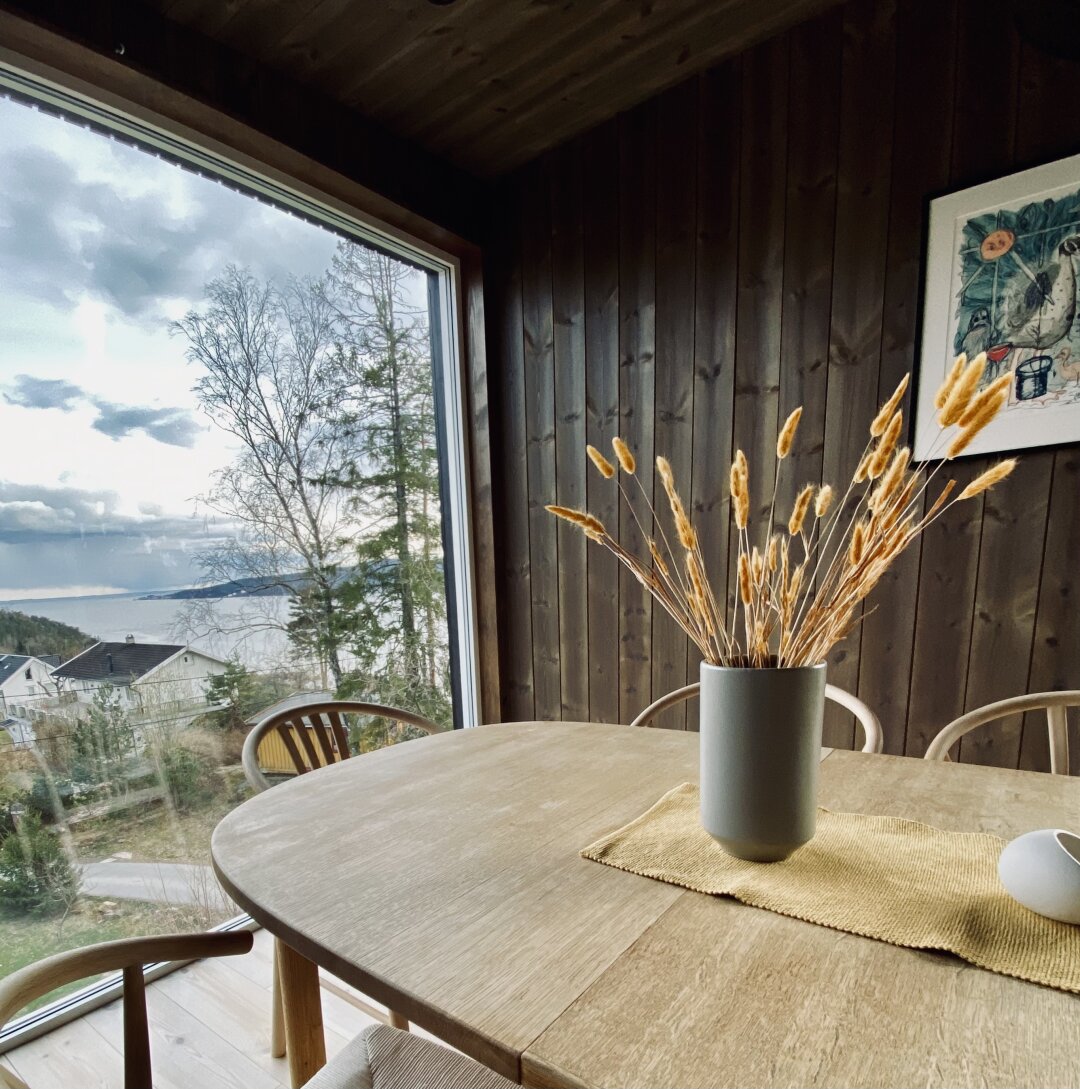 Et stort vindu åpner mot et fjordlandskap, mest et interiør av noen strå i en vase på et bord.