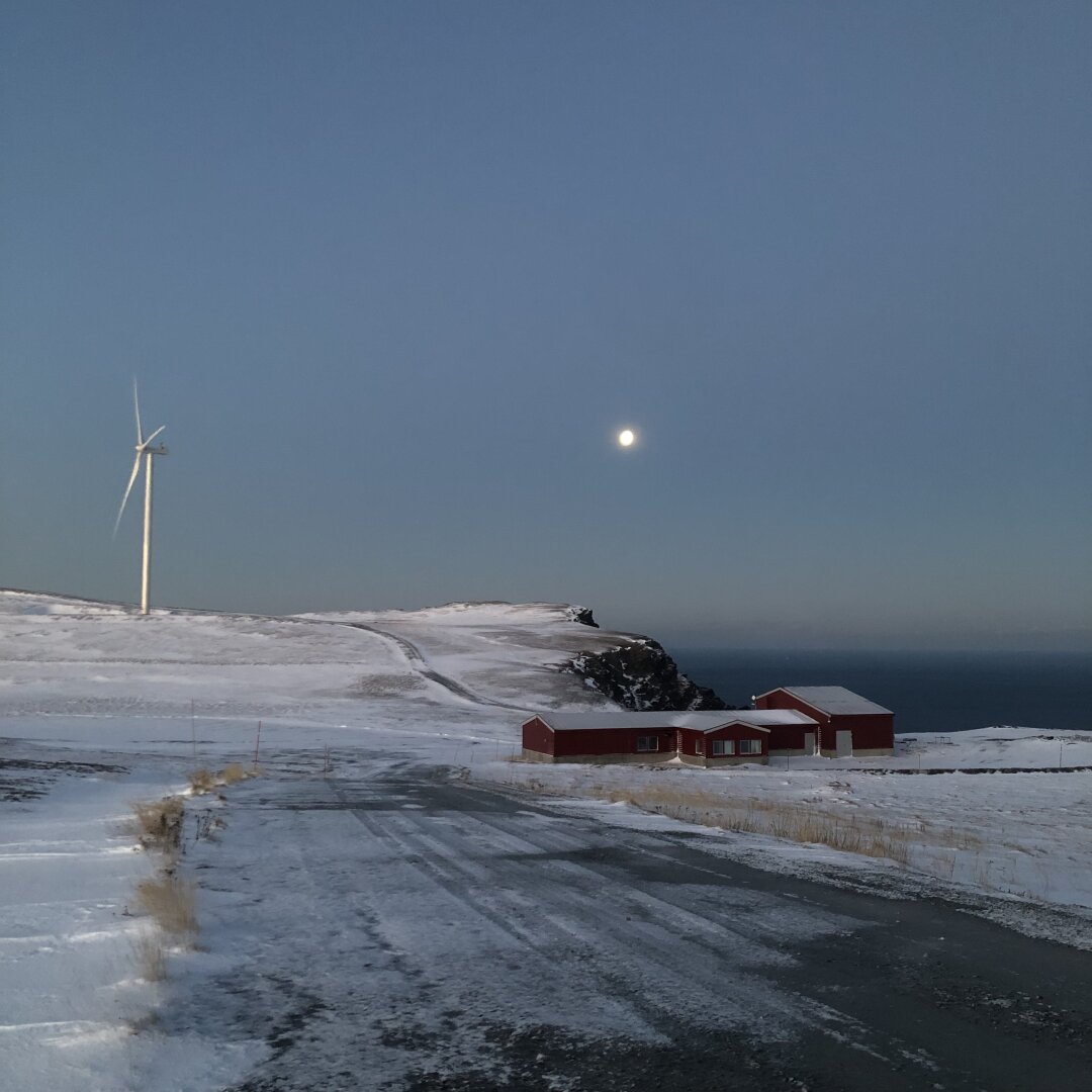 Det kan se ut som om bildet er tatt på natta, men det er faktisk bare litt seint på formiddagen. Himmelen er turkis, og det er en vei med snø, vindturbin og en måne som henger over.