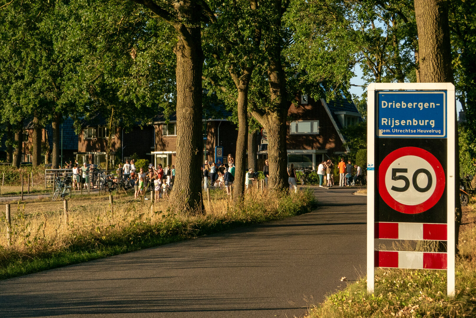 Mensen staan met eclipsbrillen en camera's naar een laag staande zon (buiten beeld) te kijken in warm licht aan de rand van een dorp. Op de voorgrond groot een bord met 'Driebergen-Rijsenburg' en daaronder een 'maximum snelheid 50 kilometer per uur' bord.