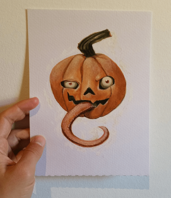 My hand holding up a paper with a goofy pumpkin with eyes and long tongue painted with oil paint.