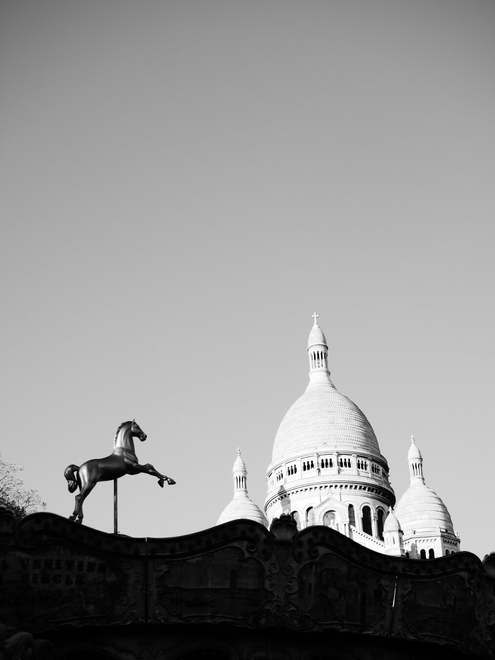 A black-and-white photograph of the top of a carousel, and just behind, the top of the Basilica of Sacré Cœur de Montmartre.