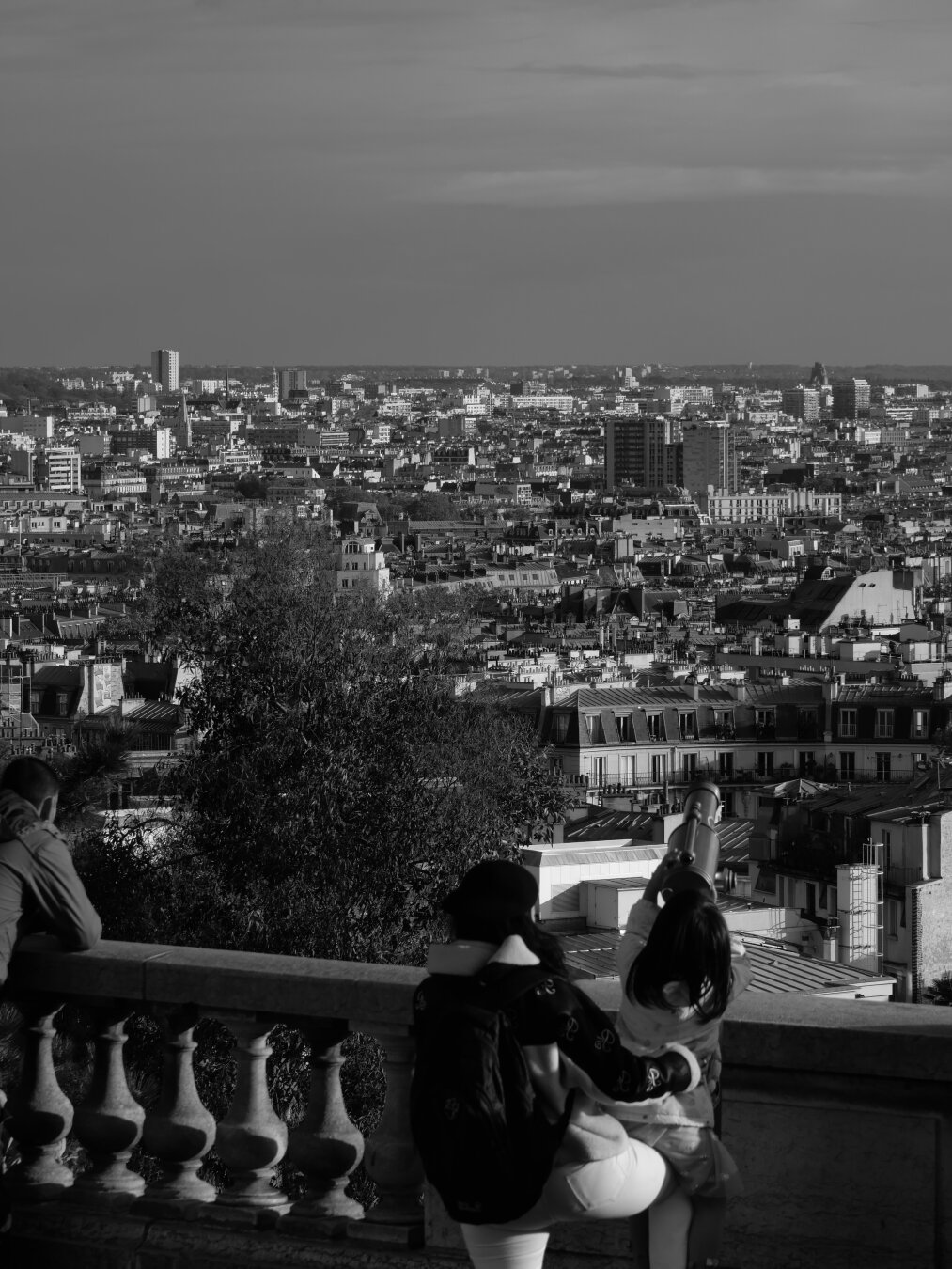 A black-and-white photograph of a girl and her mother carrying her to see Greater Paris through a spyglass. This scene takes place at the top of Montmartre hill.