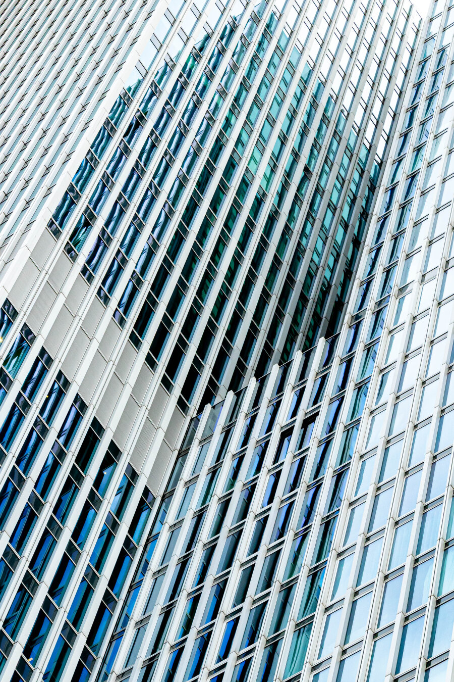 Photo of part of an office building, taken from below and diagonally (left to right). On the left is a tall, rectangular building with another similar building perpendicular to it halfway across, but half of this building is lower where it meets the first building. This creates a space between the buildings at the top. The second building is reflected in the windows of the first building. This creates a mirrored cube, bluish, likely due to the light from the sky. The buildings have rows of vertical white stripes between the rows of glass windows. Because of these rows of diagonal white stripes and the cube at the top, it's hard to see how they fit together.