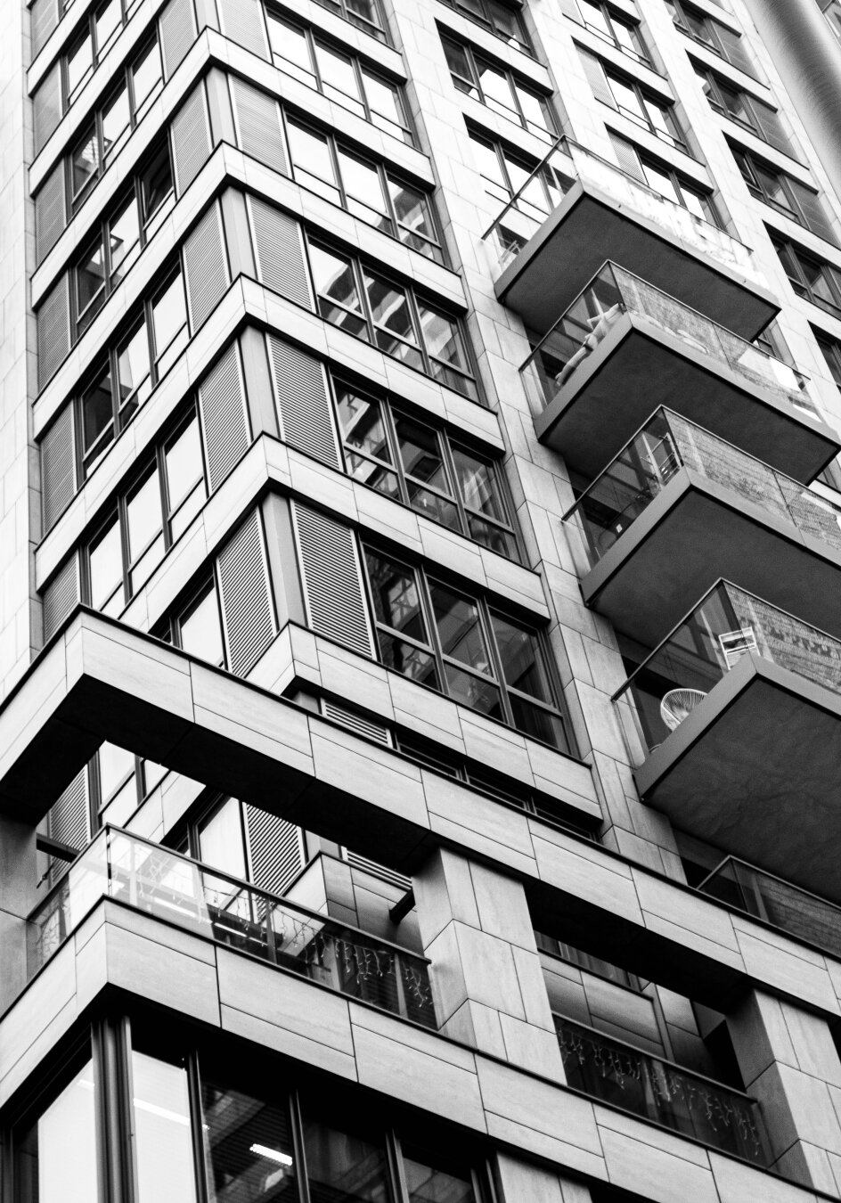 Black and white photograph of a residential tower, shot from below, slightly diagonally, at the corner of the building. At the bottom from left to right, two wide, white, L-shaped beams project forward on two angular pillars. Above them are repeating, recessed rows of windows, with V-shaped , white, sections between them. On the right is a row of balconies. Black shadows accentuate the white lines.