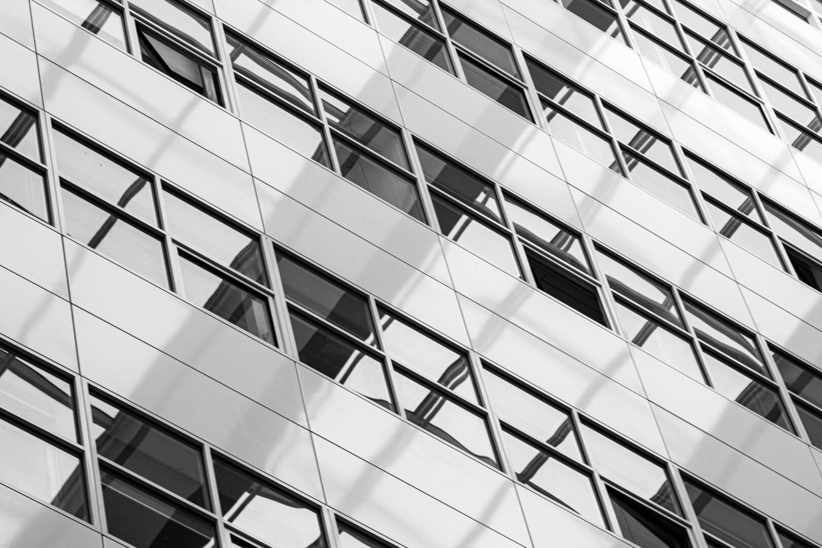 The white side wall with rows of windows of the large inner hall of The Hague City Hall was photographed from below and diagonally. The ceiling, with its large glass panes, is reflected in the windows. The wide white lines of the side wall run from left to right, and the mirrored dark gray lines of the ceiling run from left to right, crossing eachother