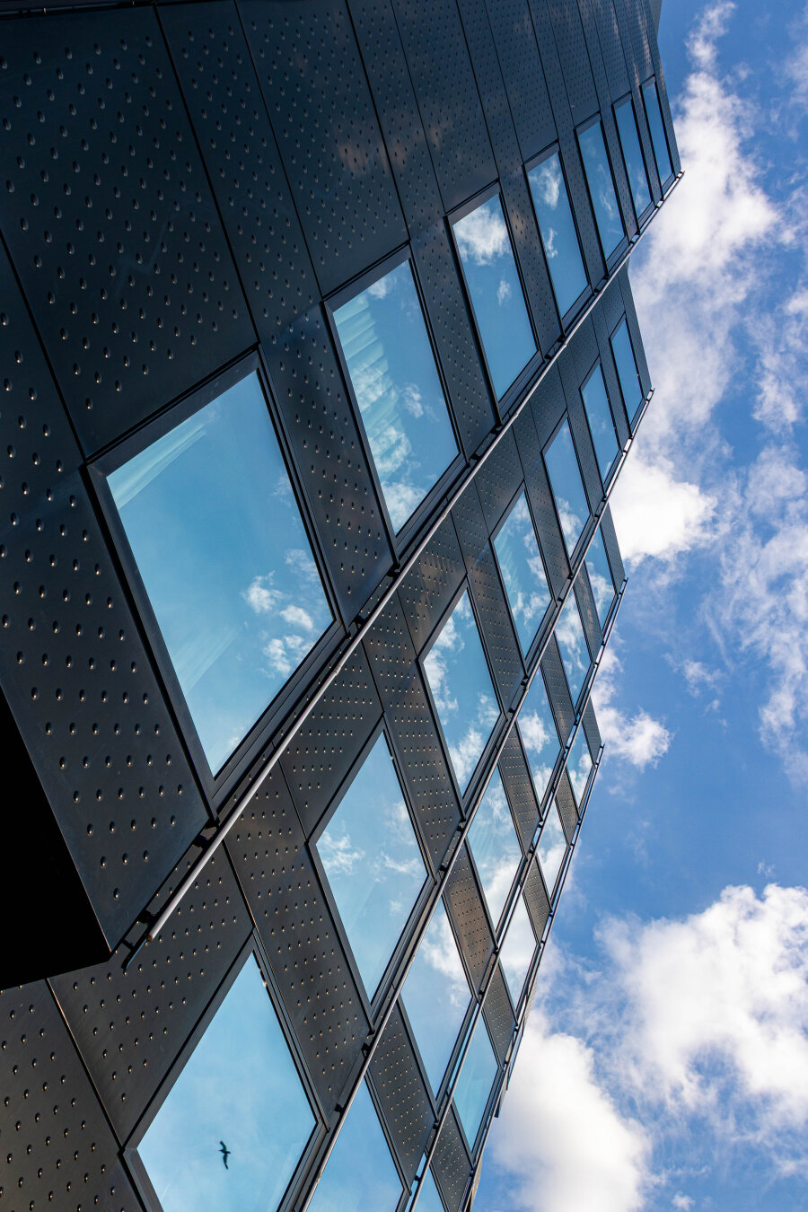 On the left, the facade of an office building, photographed from below and diagonally. To the right of the building, the blue sky and clouds stretch across its entire length. The sky and clouds are also reflected in the building's many windows. It appears as if there is only a wall with sky behind it.