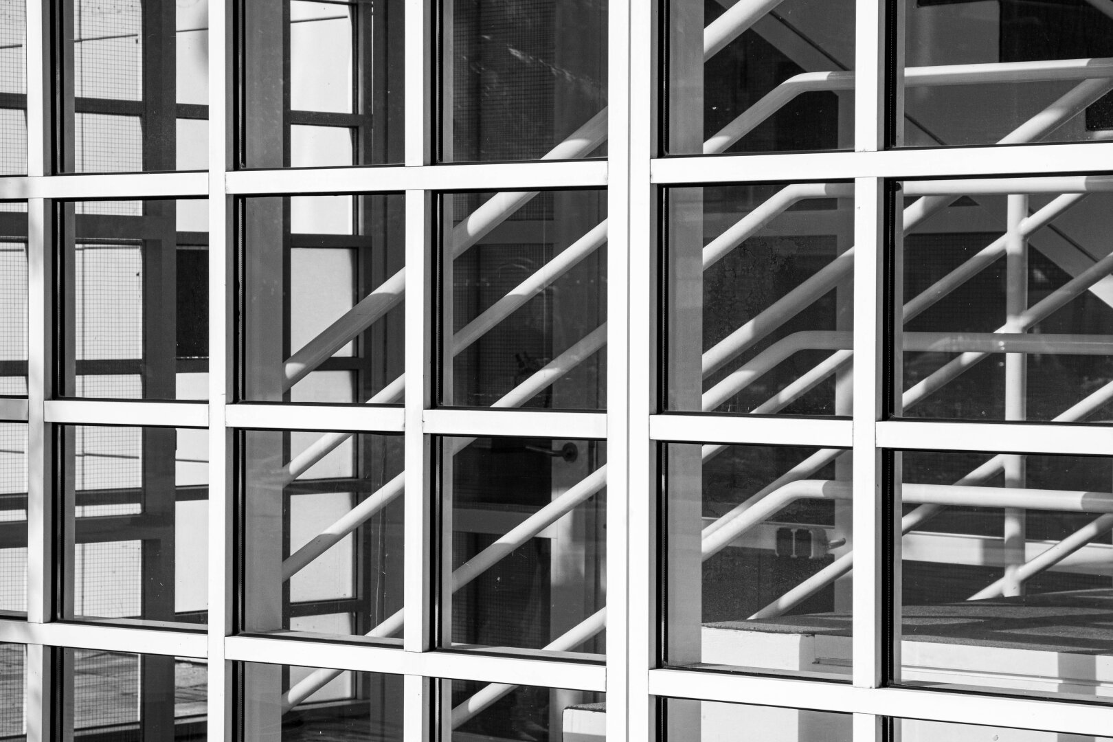 The staircase, photographed from the outside through glass, white, square panes. The lines of the white, round, and slanted banisters on the first floor intersect with horizontal and vertical white lines of the small square panes