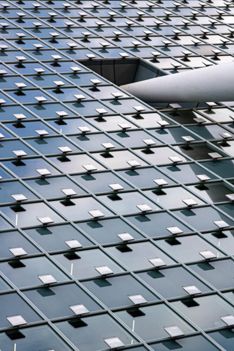 The facade of the KPN building in Rotterdam. Photographed diagonally from below. The blue sky and white clouds reflect in the glass windows. Rows of square lights on the windows are themselves reflected in the windows. In the upper right corner is a large square hole where a section of a large, slanted white pillar protrudes.