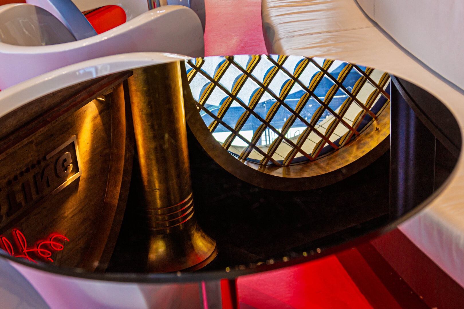 This photo is about colors and shapes that create an abstract image. It is a photo of a black glass table from above, shot diagonally in a bar. A semicircular window with checked copper bars and gold-colored wall decorations reflects in the table. Around the table are white, red and pink shades of leather chair parts.
