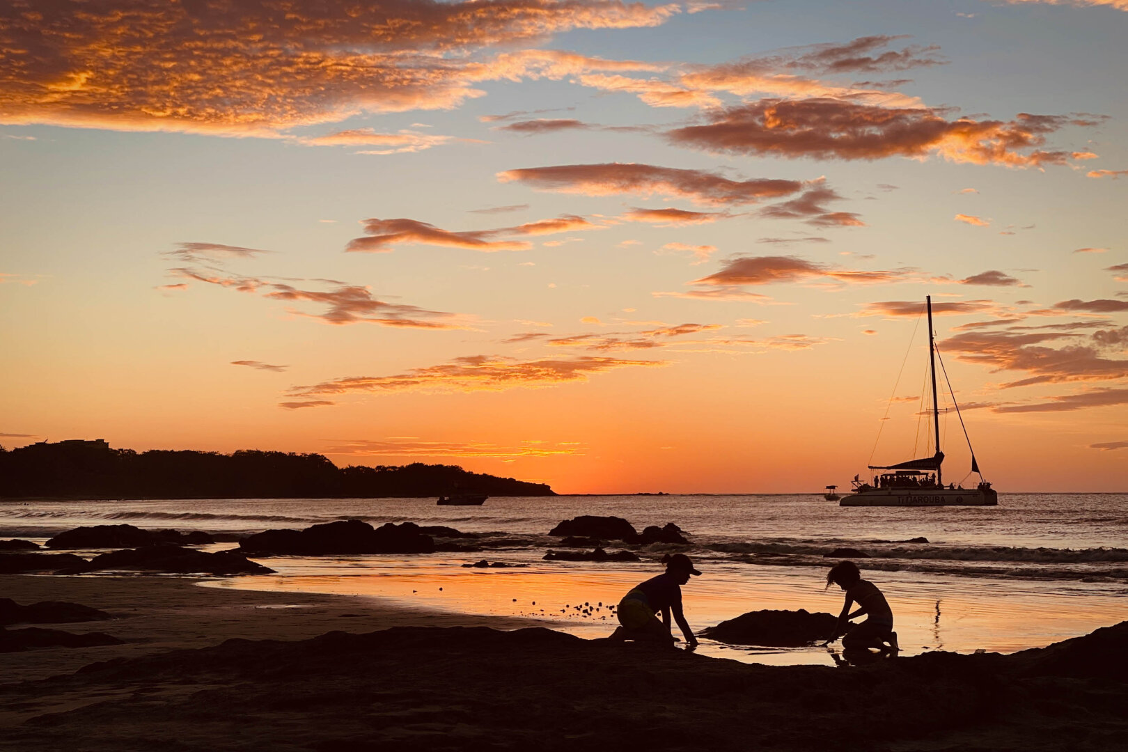A horizontal photo of two kids playing on the beach during a vibrant pink and orange sunset with the ocean looking glassy in the background. A few clouds in the sky and a boat to the right in the water behind.