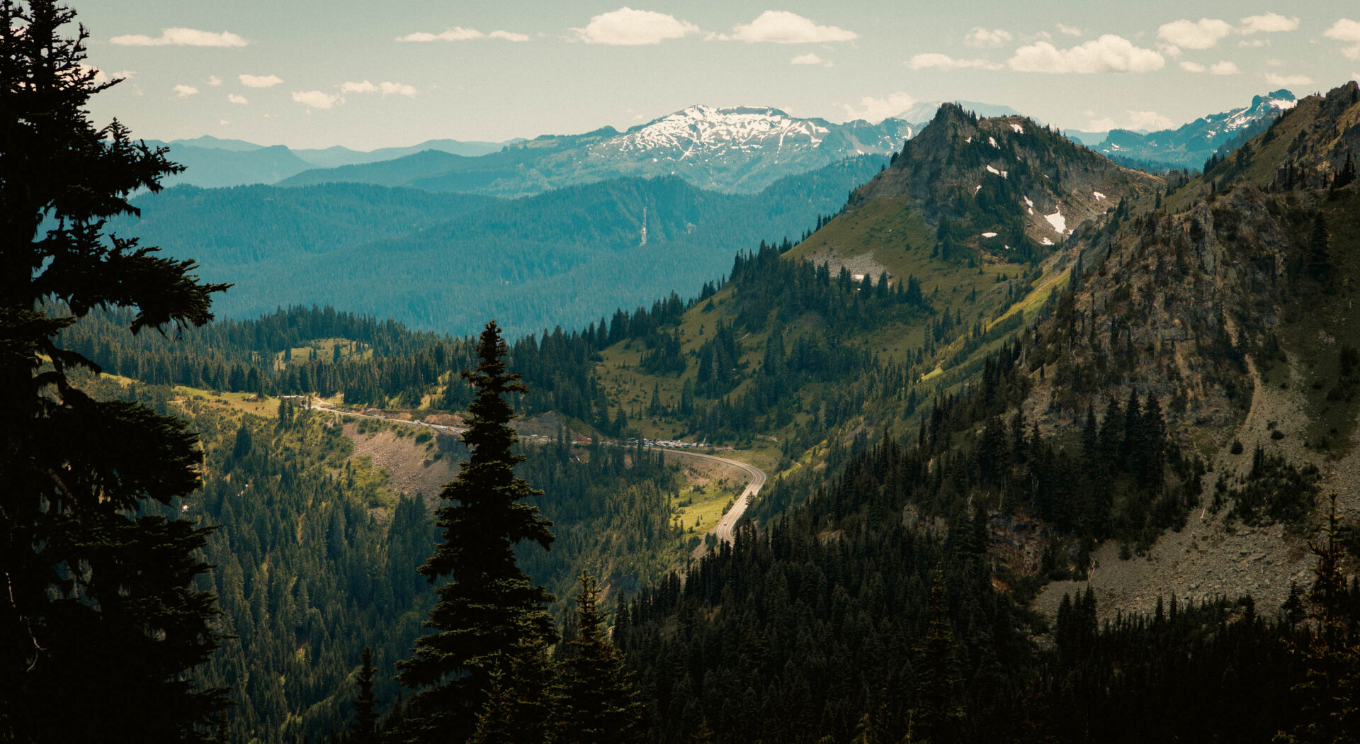A horizontal landscape overlooking a highway winding through a mountain pass with layers of blue mountains in the background and green trees in the foreground.