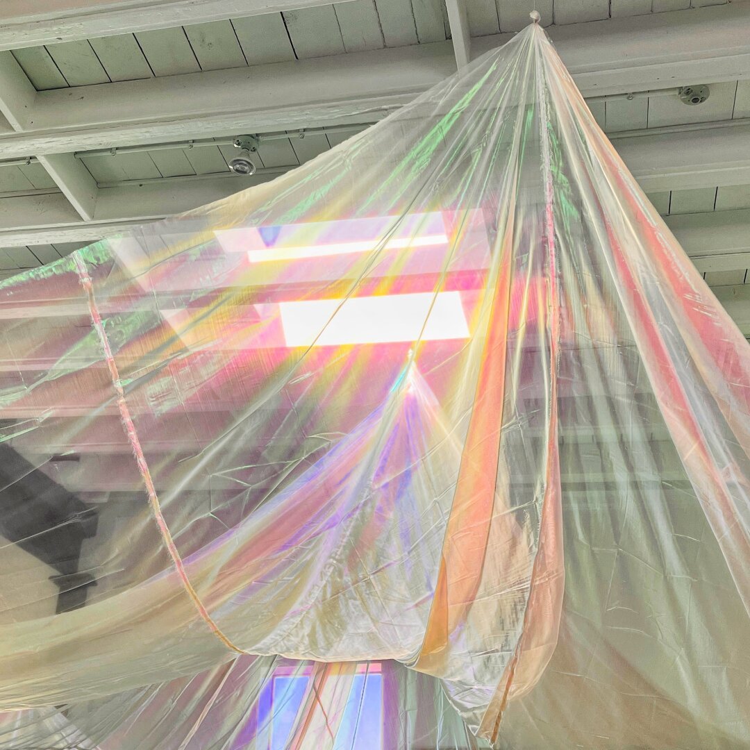 A gossamer iridescent sheet hangs from one point from a white painted wood factory ceiling. A skylight allows the sheet to show rainbow colors as it hangs
