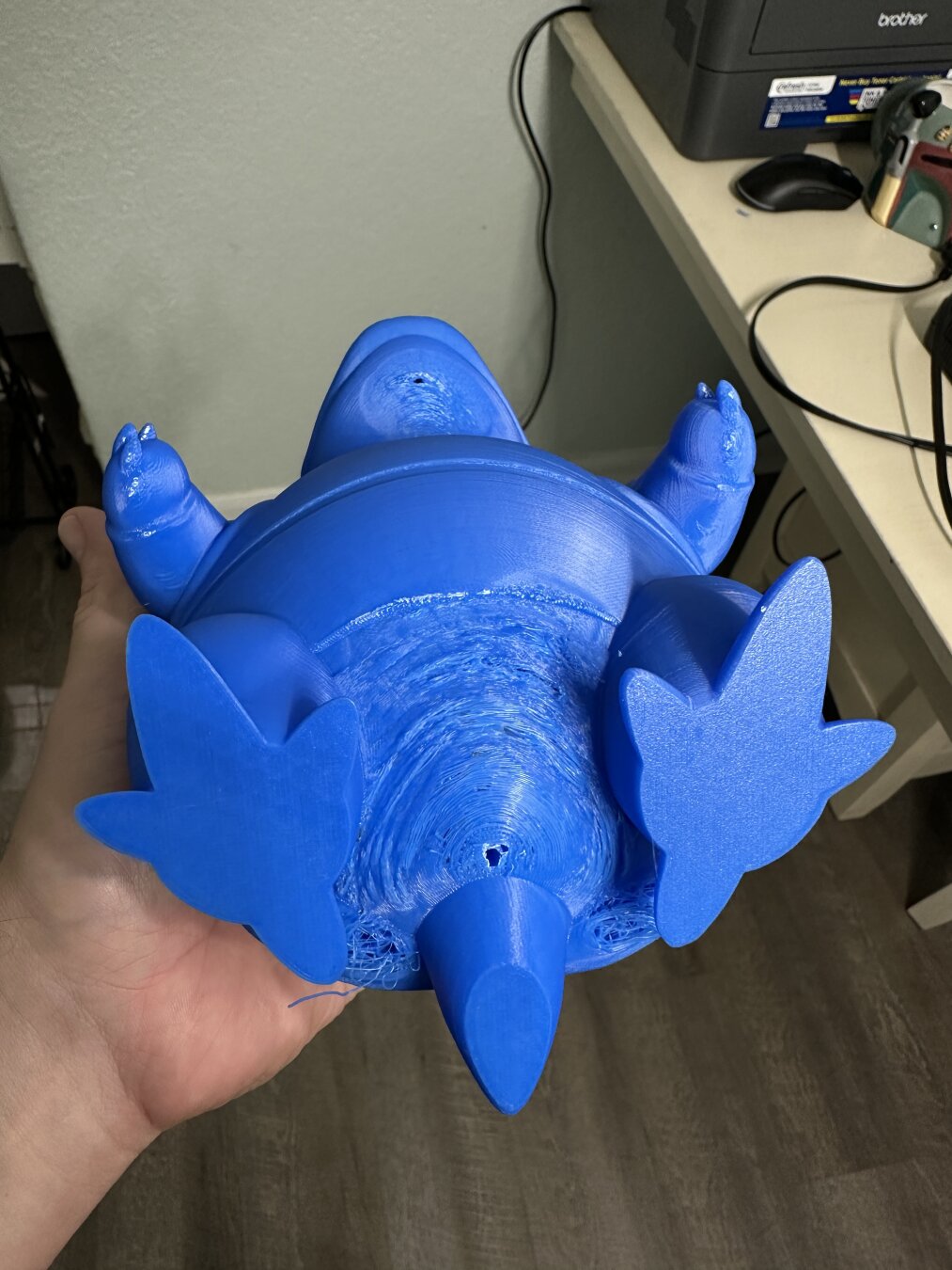 A close-up shot shows a hand holding a large, royal blue 3D-printed object, possibly a toy or figurine. The object is shaped like a cartoon turtle or tortoise, lying on its back, revealing its underside. It has four flipper-like feet, a rounded body, and a small, pointed tail. The surface of the object shows visible layers from the 3D printing process, particularly on the underside, which has a somewhat rough, textured appearance. In the background, out of focus, parts of a room are visible, including a light-colored desk or table with a printer and some cables.