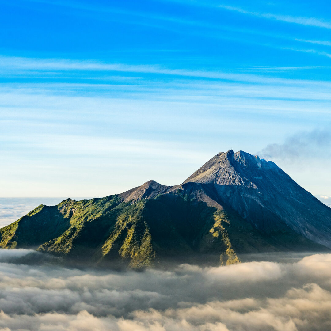 Photo of a volcano with clouds at the base of the mountain, with slight smoke from the peak.