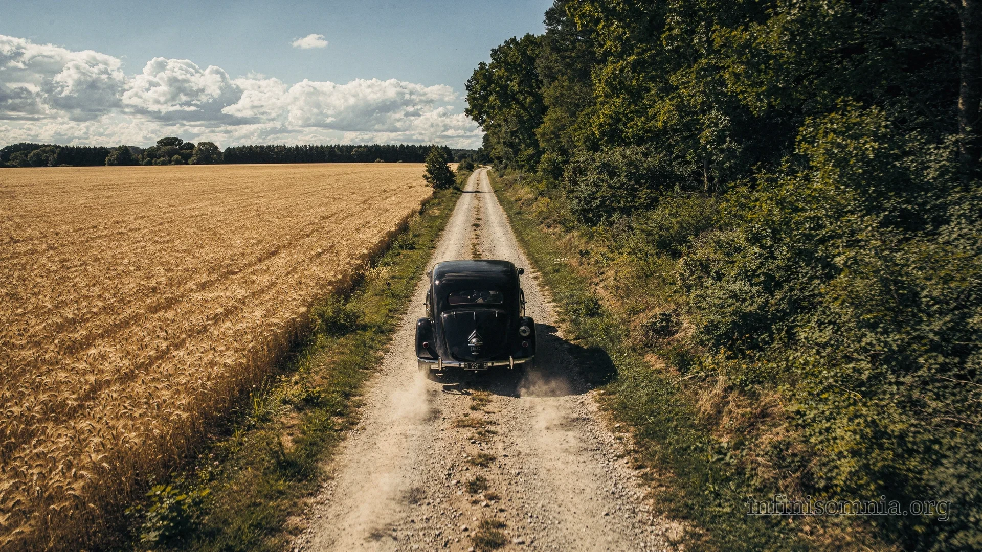 A black vintage car is driving along a country road between a field and a forest. The weather is summery and pleasant, but storm clouds are already gathering in the distance.

The image is similar to the previous one, but looks a lot more realistic. Also the car is now in a good proportion to the street and wheat field.