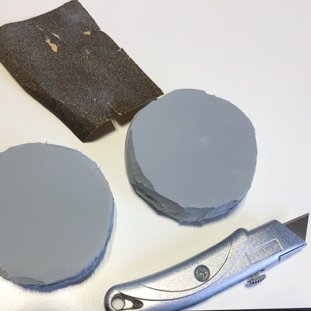 Process of crafting a LARP throwing weapon: Two roughly cut stacks of circles from grey EVA foam glued together to create the base of the cinnamon bun prop. Next to them lies a cutting knife and a piexe of sandpaper.