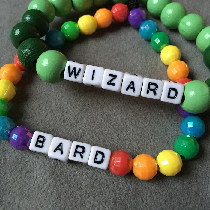 Two handmade beaded bracelets displayed on grey fabric: one rainbow-coloured with white letter beads spelling “BARD”, the other in green tones with the word “WIZARD”. Both are custom jewellery pieces inspired by tabletop role-playing games like Dungeons & Dragons. Ideal for expressing your character’s class or identity.