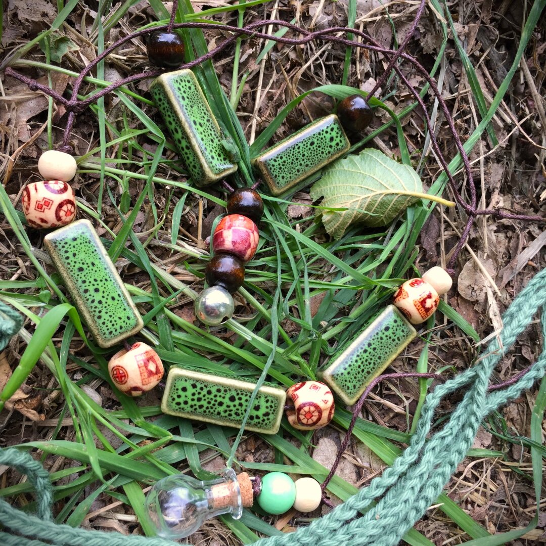 Several pieces of handmade jewelry are placed in the green grass among some leaves. Two necklaces are made from brown and beige wooden beads and glazed rectangular parts in forest green on a dark brown string. Another bead has a tiny potion bottle attached to it, and there is a crocheted green hair tie next to it.
