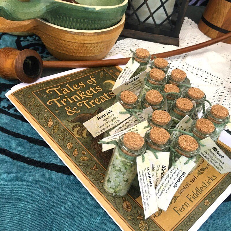 A collection of small bottles with Forest Salt, a greenish spiced salt with  wild garlic and young spruce and pine tips. It is pictured on a table with a cottage tablecloth, a long wooden smoking pipe, and a printed collection of tales by Fern Fiddlesticks, a travelling hobbit.