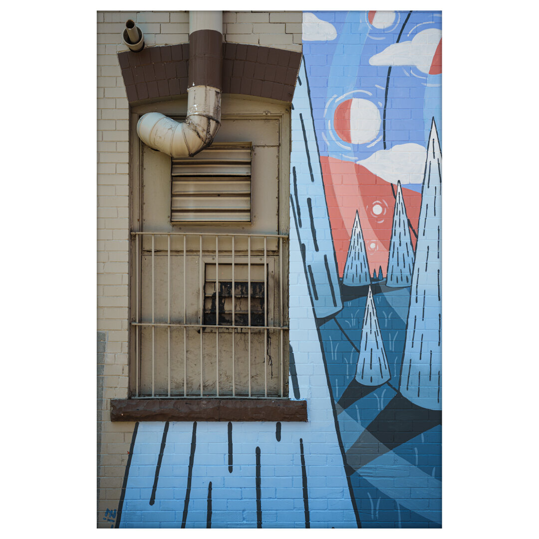 A photograph of a building exterior featuring a heavily weathered and barred window with a rusted exhaust fan behind metal bars. Above the window, an old ventilation pipe curves outward and up, attached to a beige wall made of painted bricks. The right side of the wall is covered in a vibrant mural: a stylized landscape with triangular trees, flowing lines, floating clouds, and large red and white circular shapes resembling suns or moons. The mural's palette includes sky blue, navy, coral red, and white, contrasting sharply with the dull, industrial elements of the window and pipe. The image captures the tension between decay and creativity in an urban setting. A photograph of a building exterior featuring a heavily weathered and barred window with a rusted exhaust fan behind metal bars. Above the window, an old ventilation pipe curves outward and up, attached to a beige wall made of painted bricks. The right side of the wall is covered in a vibrant mural: a stylized landscape with triangular trees, flowing lines, floating clouds, and large red and white circular shapes resembling suns or moons. The mural's palette includes sky blue, navy, coral red, and white, contrasting sharply with the dull, industrial elements of the window and pipe. The image captures the tension between decay and creativity in an urban setting.