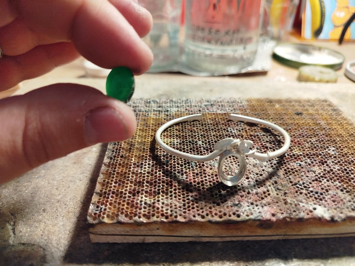 a hand holds an oval green gemstone. a silver bracelet in the background which is in progress of making it