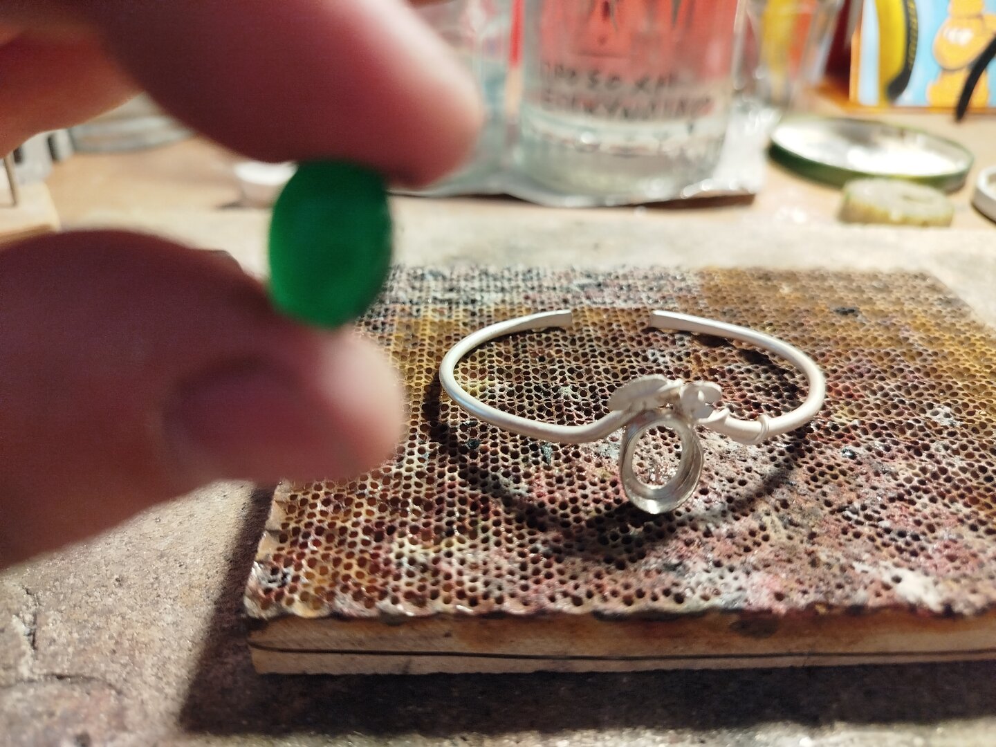 a hand holds an oval green gemstone. a silver bracelet in the background which is in progress of making it