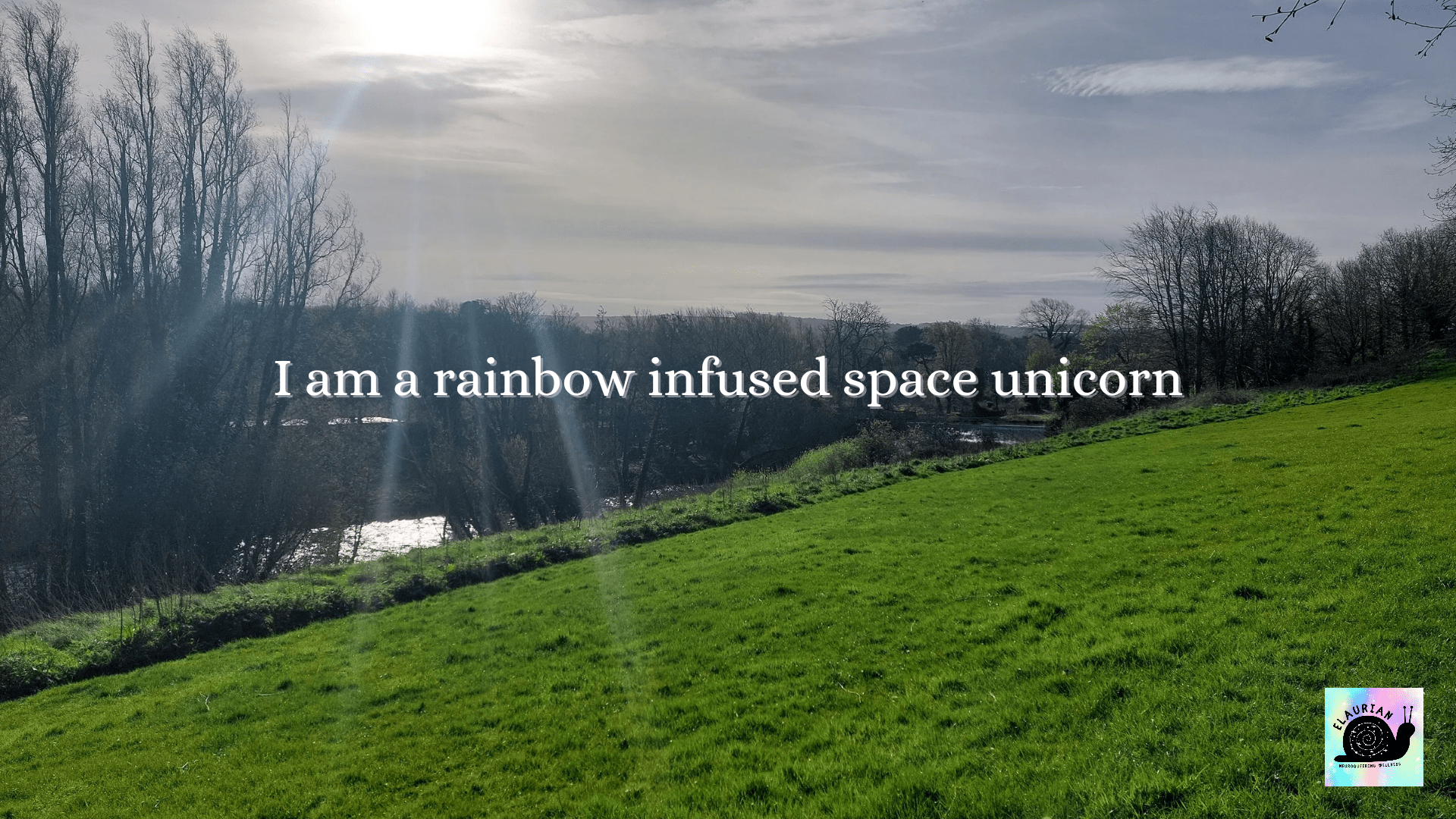 A sloping hill with a river and bare trees at the bottom. The sun is shining down over all of it, and in white writing it says: I am a rainbow infused space unicorn.