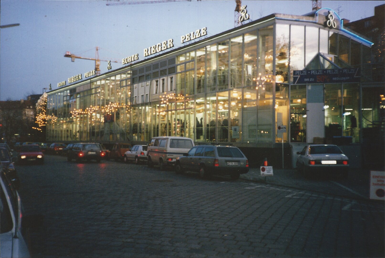 A low-light, street-level photograph of a large, modern commercial building with a glass facade, prominently featuring the illuminated sign "ECHTE RIEGER PELZE" (Real Rieger Furs). The building's interior is brightly lit, and the street in the foreground is cobblestone with several parked cars.